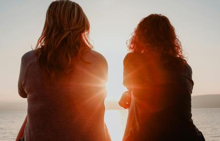 Illustrasjonsbilde. To jenter sitter med ryggen til, og ser mot solnedgang som går ned i havet.