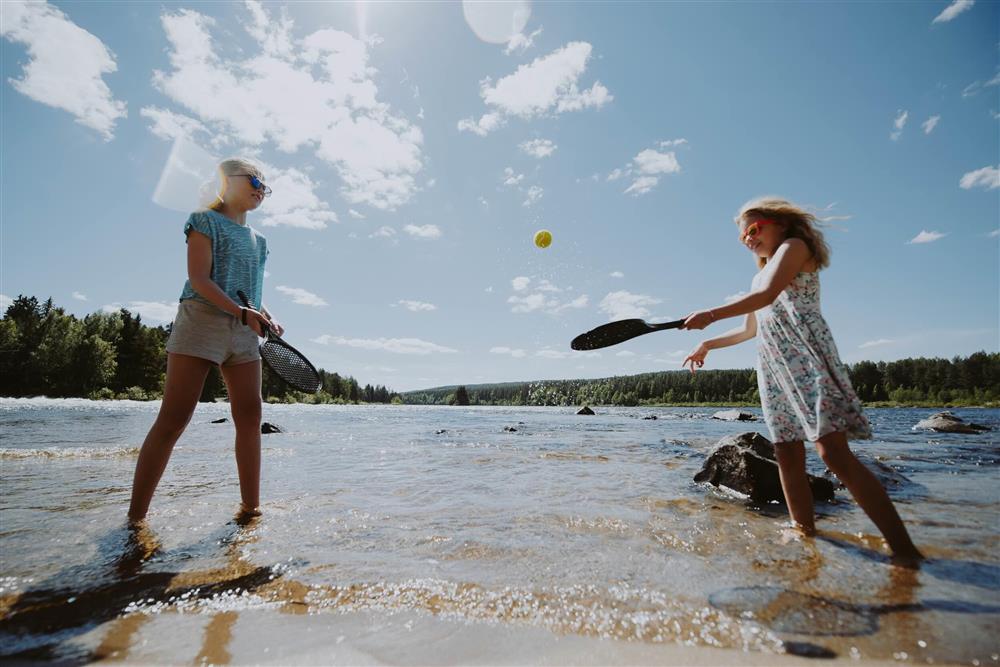 To jenter som spiller badminton på en strand ved en elv. - Klikk for stort bilde