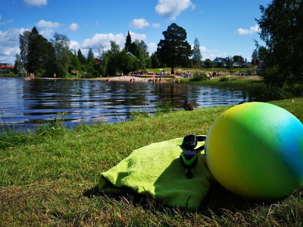Et badehåndkle og en ball ligger på gress med et badetjern. I bakgrunnen en strand med mange folk. - Klikk for stort bilde