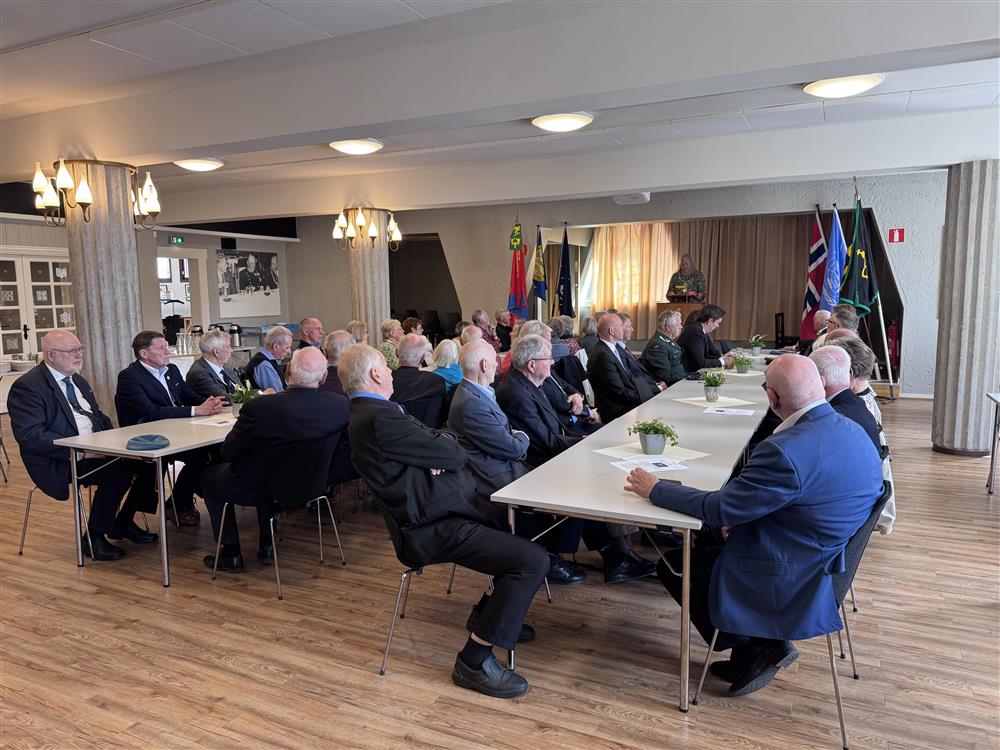 Mange personer sitter rundt to langbord og ser mot en mann som taler fra en liten scene. - Klikk for stort bilde